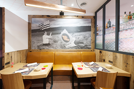 Deux tables situées derrière le bar. La banquette en cuir orange est surplombée par une illustration en noir et blanc d'un homme portant fièrement le drapeau breton. On peut aussi y voir le logo du restaurant. 