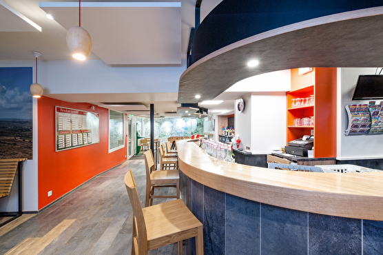 Le comptoir de bar est arrondis et de couleur bleu, les chaises hautes sont en bois clair et certains murs du Café sont peints en orange